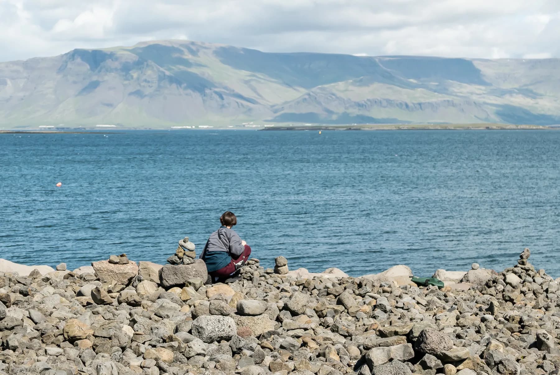 Person sitting on a rocky shoreline in Reykjavik, looking out across blue water toward distant mountains.