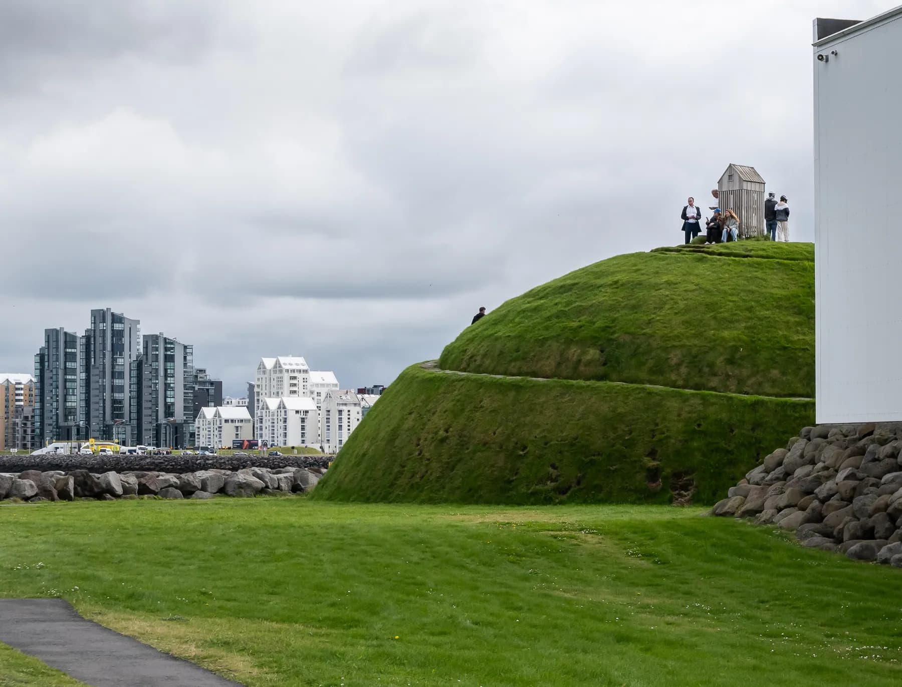 Grassy hill and stone wall in Reykjavik with modern buildings visible in the distance under a cloudy sky.